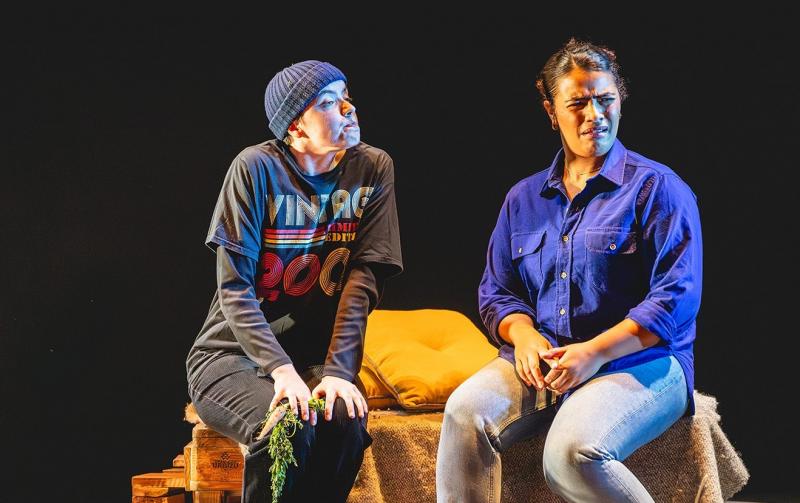 Two young actors on stage sit side-by-side on a bench